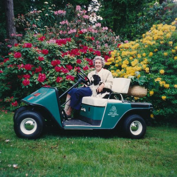 Woman and a small dog sitting on a 2-seater golf cart in front of colourful, blooming rhododendrons