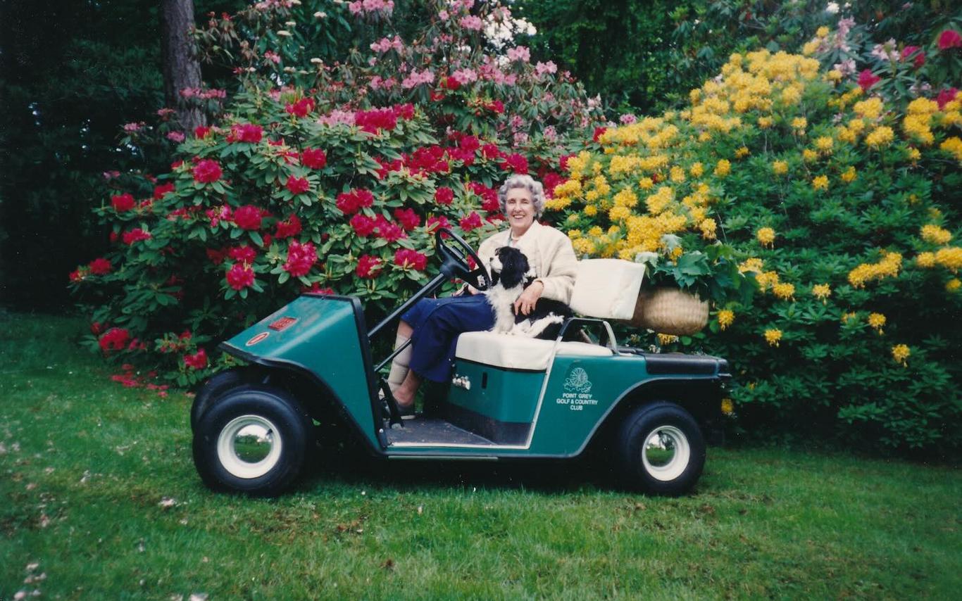 Woman and a small dog sitting on a 2-seater golf cart in front of colourful, blooming rhododendrons